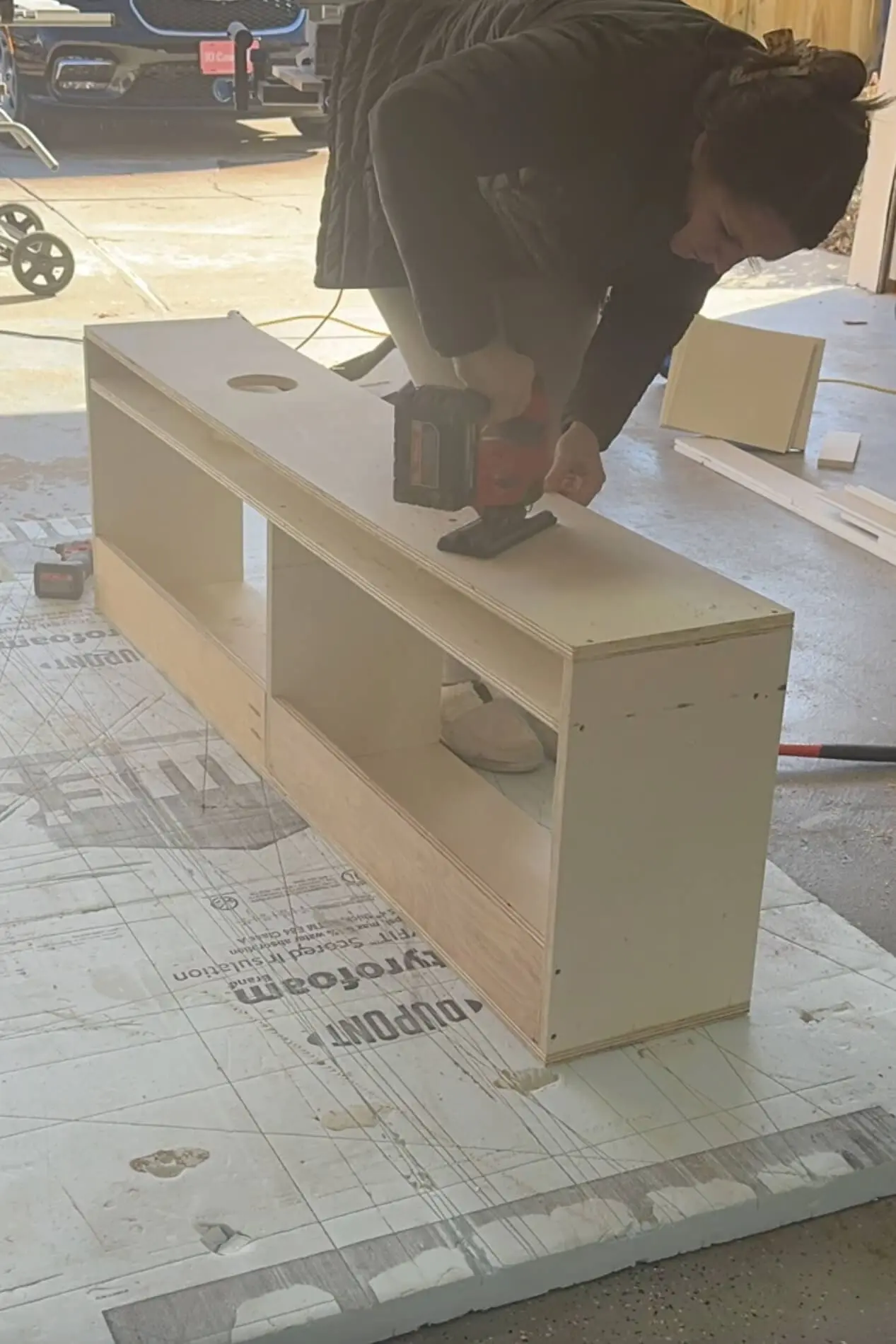 A woman sanding the top surface of a DIY wooden storage bench using a power sander. The structure is fully assembled with open storage compartments, and a circular hole has been cut into the top. She is wearing a green puffer jacket and fuzzy slippers, focusing on smoothing out the wood for a polished finish.